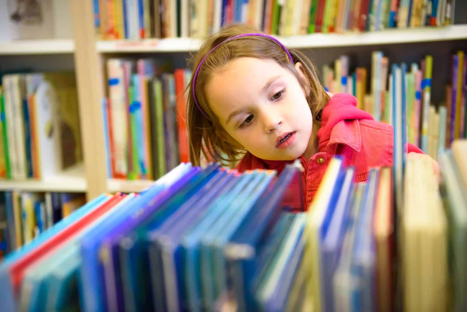 Little girl is choosing a book in the library. A child is looking at the books in the library deciding which one to take home. Children creativity and imagination.